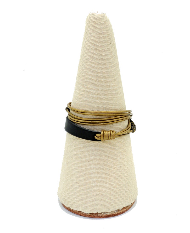 May include: A handcrafted ring with a black leather band and gold-toned wire detailing. The ring is displayed on a white cone-shaped stand, highlighting its unique design. The ring's design incorporates a mix of materials and textures, showcasing a modern aesthetic.