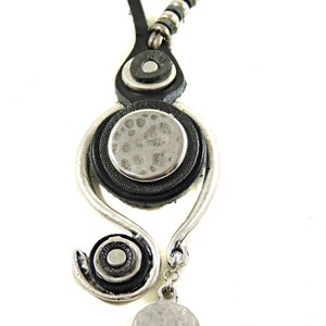 Pu&ograve; includere: Una collana in pelle nera con un ciondolo d'argento che presenta un cerchio con una superficie testurizzata e un cerchio pi&ugrave; piccolo con una superficie liscia. La collana ha una catena d'argento con un ciondolo rotondo d'argento.