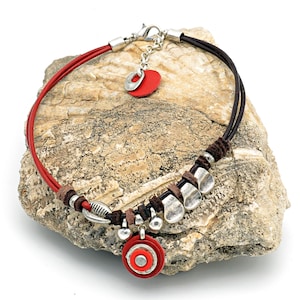Op de afbeelding: Een rode en bruine leren ketting met zilveren metalen accenten. De ketting heeft een grote rode ronde hanger met een kleinere rode cirkel in het midden. De ketting wordt gepresenteerd op een rots.