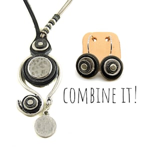 Pu&ograve; includere: Una collana in pelle nera con dettagli argentati e un paio di orecchini abbinati. La collana presenta un ciondolo rotondo con una finitura argentata martellata e gli orecchini hanno un design simile. Il testo "COMBINE IT!" &egrave; scritto sotto gli orecchini.