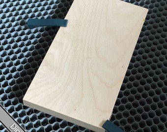 May include: A light wood board with a smooth surface, held in place by two dark blue clamps on a gray honeycomb surface.  Digital Download.