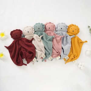 Pode incluir: Seis mantas de segurança de pelúcia em forma de leão em diferentes cores: bordô, bege, azul-petróleo, rosa, cinza e amarelo. Cada manta apresenta um rosto de leão com olhos pretos e um nariz preto.