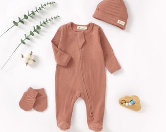 May include: A three-piece baby outfit set in a dusty rose color. The set includes a long-sleeved ribbed onesie with a zipper closure, a matching ribbed hat, and a pair of ribbed mittens.