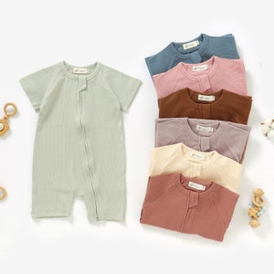 May include: A set of seven baby onesies in various colors, including green, pink, brown, beige, and blue. The onesies are made of a ribbed fabric and have a zipper closure.