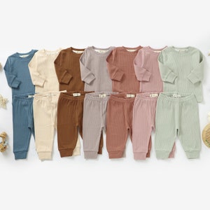 May include: Ten sets of baby pajamas in various colors, including blue, beige, brown, purple, pink, and green. Each set includes a long-sleeved shirt and matching pants.