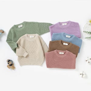 May include: A collection of six children's sweaters in various colors: sage green, beige, lavender, light blue, brown, and dusty rose. Each sweater has a ribbed knit texture and a crew neck. The sweaters are arranged on a white surface.