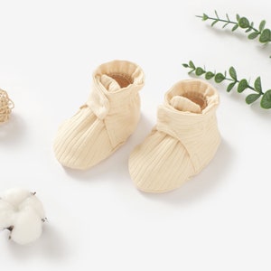 May include: A pair of cream-coloured baby booties with a ribbed texture and a button detail. The booties are made of soft, comfortable fabric and are perfect for keeping little feet warm.
