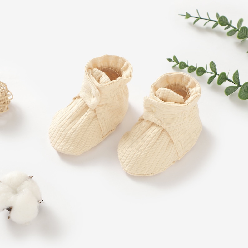 May include: A pair of cream-coloured baby booties with a ribbed texture and a button detail. The booties are made of soft, comfortable fabric and are perfect for keeping little feet warm.