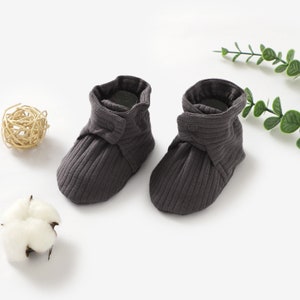 May include: A pair of grey baby booties with a button fastening. The booties are made from a soft, ribbed fabric.