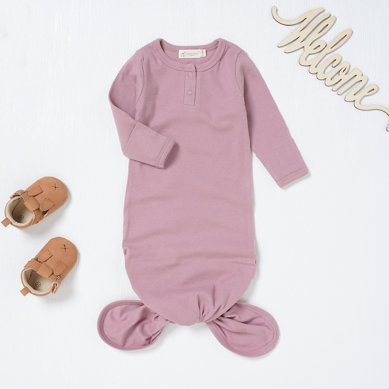 May include: A mauve baby sleep sack with long sleeves and a knotted bottom. The sleep sack has a single button closure at the neckline.