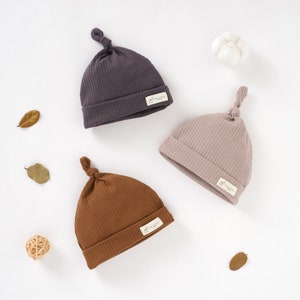 Può includere: Tre cappellini per neonati in diversi colori: marrone, grigio e rosa. Ogni cappellino ha un nodo sulla parte superiore e una piccola etichetta con il testo "Tiny & Wild" sul lato.