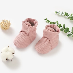 May include: A pair of pink baby booties with a button fastening. The booties are made from a soft, ribbed fabric.