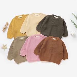 May include: Six different coloured knitted sweaters for babies or toddlers. The sweaters are in shades of brown, pink, yellow, beige, and green.