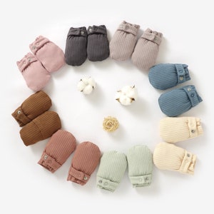 May include: A set of 10 pairs of baby mittens in various colours, including brown, pink, beige, blue, and green. The mittens are made of soft, ribbed fabric and have a button fastening.