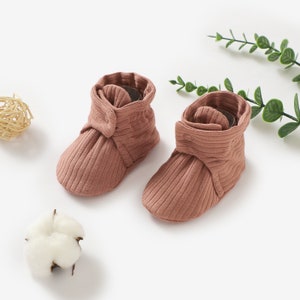 May include: A pair of brown ribbed baby booties with a button detail. The booties are made of soft, stretchy fabric and are perfect for keeping little feet warm and cosy.