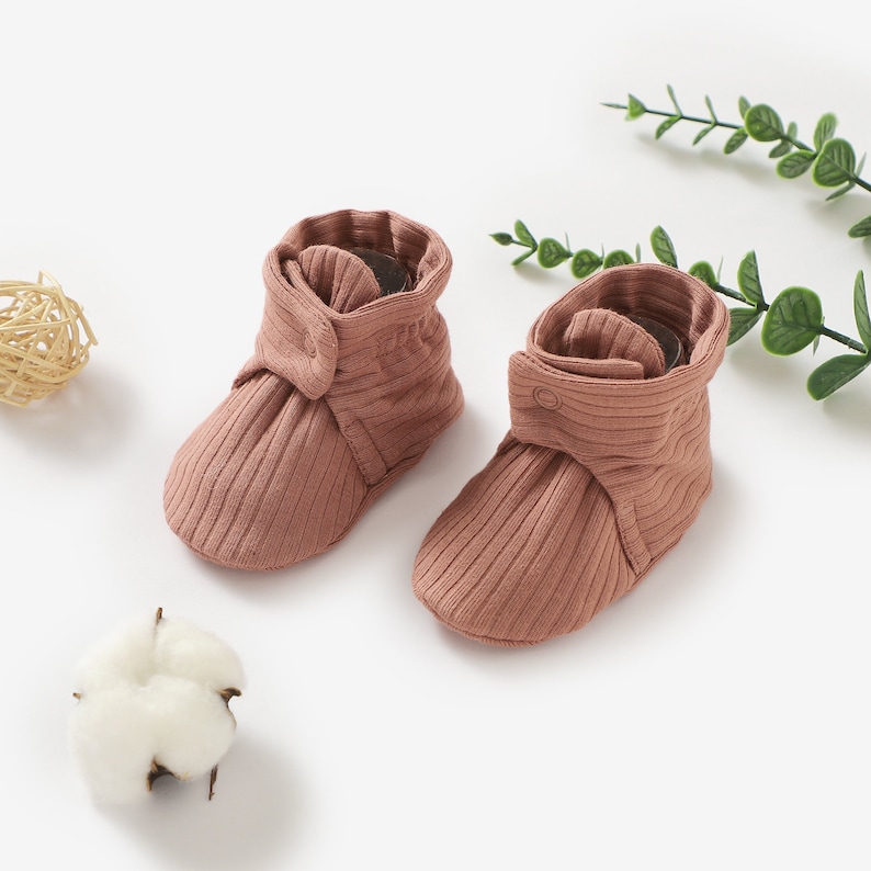 May include: A pair of brown ribbed baby booties with a button detail. The booties are made of soft, stretchy fabric and are perfect for keeping little feet warm and cosy.