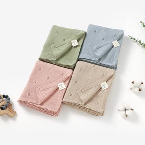 May include: Four folded baby blankets in pastel colors: green, blue, pink, and beige. Each blanket has a delicate knit pattern with small holes and a tag. The blankets are arranged on a white surface, with cotton buds and greenery.