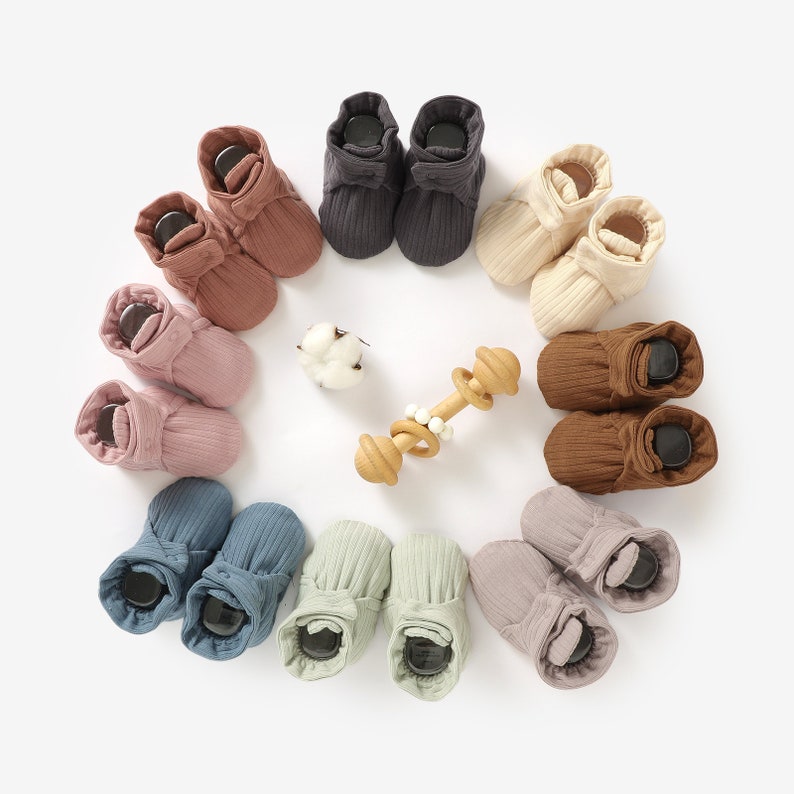 May include: A set of 10 pairs of baby booties in various colours, including brown, beige, grey, blue, and green. The booties are made of a soft, ribbed fabric and have a fold-over cuff.