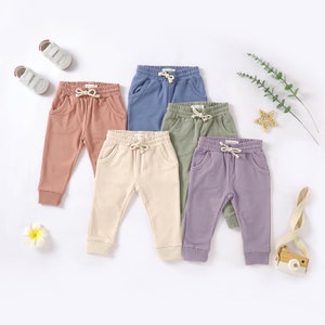 May include: Five pairs of baby sweatpants in different colours: light pink, beige, light green, light blue, and purple. The sweatpants have an elastic waistband and a drawstring.