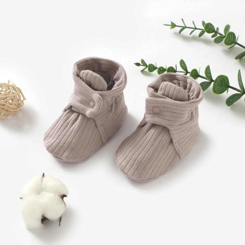 May include: A pair of soft, grey baby booties with a ribbed texture and a button closure. The booties are made of a comfortable, breathable fabric.