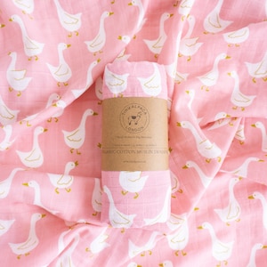 Könnte beinhalten: Rosa Bio-Baumwoll-Musselin-Swaddle mit einem weißen Gänsemuster mit goldenen Akzenten. Die Swaddle ist aufgerollt und mit einem braunen Papierband mit dem Text "Tiny Alpaca London" und "Organic Cotton Muslin Swaddle" umwickelt. Die Swaddle misst 106 cm x 120 cm.