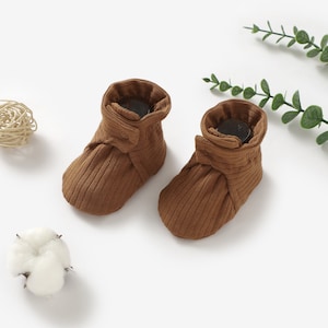May include: A pair of brown baby booties with a ribbed texture and a button detail. The booties are made of soft, comfortable fabric and are perfect for keeping little feet warm.