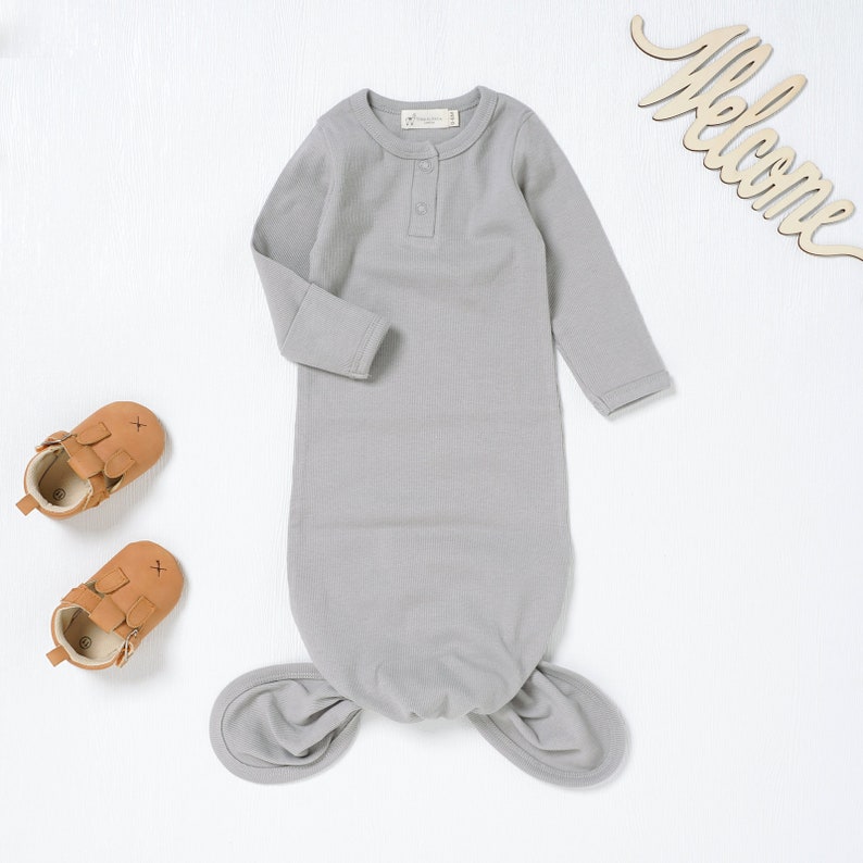 May include: A gray baby knotted gown with long sleeves and a single button closure at the neckline. The gown has a knotted design at the bottom.