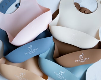 Puede incluir: Varios baberos de silicona suave para bebés en colores pastel, como rosa, azul, beige y marrón. Cada babero tiene un bolsillo curvo y el texto "TINY ALPACA LONDON".