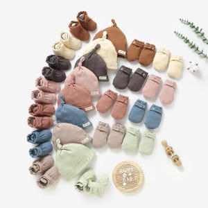May include: A set of baby hats and booties in various colors, including brown, beige, blue, and green. The hats are knitted and have a knotted top. The booties are also knitted and have a ribbed texture. The set is arranged on a white background with a wooden sign that says "Happy Baby".