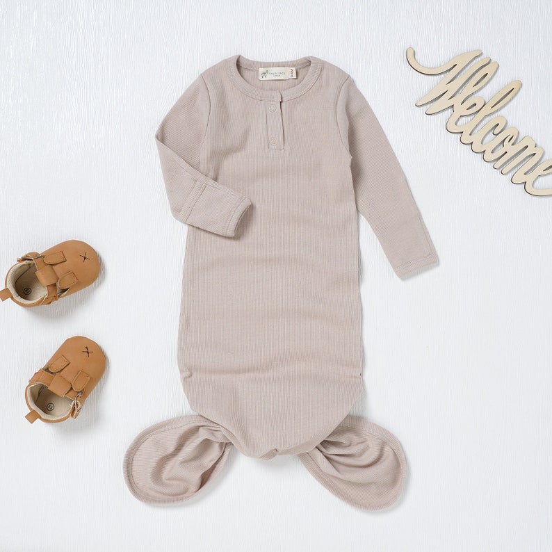 May include: A light brown baby sleeping bag with a knotted bottom and long sleeves. The sleeping bag has a single button closure at the neckline.