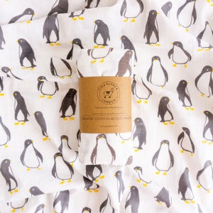 Puede incluir: Una manta de muselina de algodón orgánico blanca con estampado de pingüinos. La manta está doblada y tiene una etiqueta marrón que dice "Tiny Alpaca London" y "Organic Cotton Muslin Swaddle".