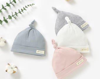 May include: Four baby hats in different colors: gray, light blue, white, and pink. Each hat has a knot on top and a small label with the text "Tiny Alpaca" on the side.