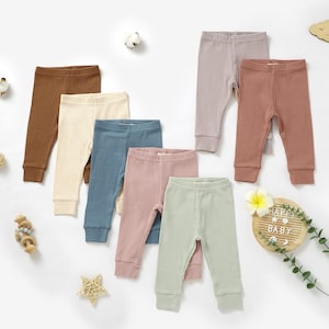 Puede incluir: Siete pares de leggings de bebé de canalé en varios colores, incluyendo marrón, beige, azul, rosa y verde. Los leggings están dispuestos sobre una superficie blanca.