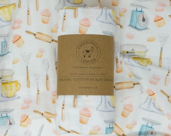 May include: A white muslin swaddle with a repeating pattern of kitchen tools and cupcakes. The swaddle is folded and has a brown label with the text "Tiny Alpaca London" and "Small Moments. Big Memories".
