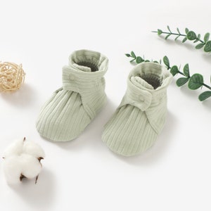 May include: A pair of light green baby booties with a ribbed texture and a button fastening. The booties are made from soft, comfortable fabric and are ideal for keeping little feet warm.