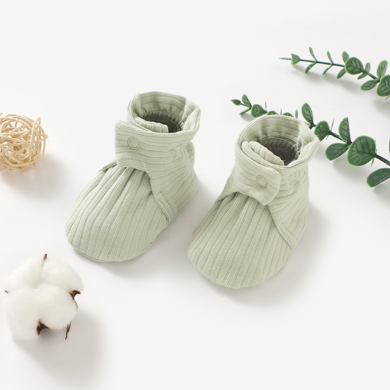 May include: A pair of light green baby booties with a ribbed texture and a button fastening. The booties are made from soft, comfortable fabric and are ideal for keeping little feet warm.