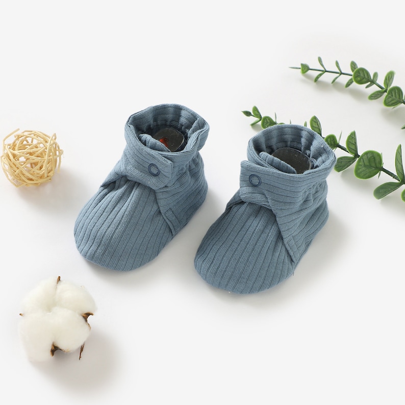 May include: A pair of blue baby booties with a button fastening. The booties are made from a soft, ribbed fabric.