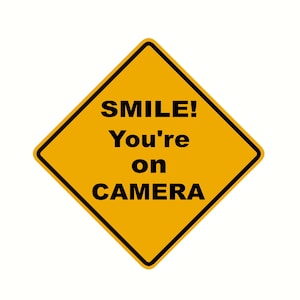 May include: A yellow diamond-shaped road sign with black text that reads "SMILE! You're on CAMERA".