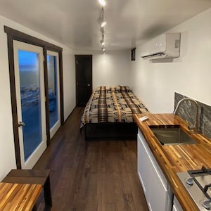 Puede incluir: Vista interior de un espacio habitable pequeño y moderno con cama, cocina y suelos de madera. La cama tiene una manta estampada. La cocina cuenta con encimera de madera, fregadero y estufa. La iluminación de riel ilumina la habitación.