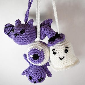 May include: Crocheted Halloween ornaments in purple and white. The set includes a bat, an eyeball, a ghost wearing a witch hat. Each ornament has black button eyes and a white string for hanging. The bat has black wings.