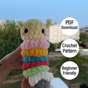 May include: A colorful, handmade crochet doll with a yellow head, black eyes, and pink cheeks. The doll's body is made of stacked rings in various colors, including pink, yellow, blue, red, green, and white. The image also includes text that says "PDF Download", "Crochet Pattern", and "Beginner Friendly".