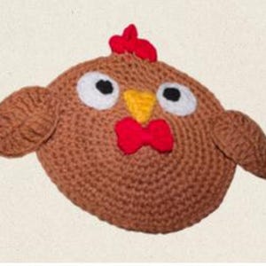 Puede incluir: Una cabeza de pollo de ganchillo hecha a mano, redonda y marr&oacute;n, con cresta y barbilla rojas, pico amarillo y ojos negros con borde blanco. El pollo tiene peque&ntilde;as alas redondeadas a cada lado. El fondo es de color beige claro.