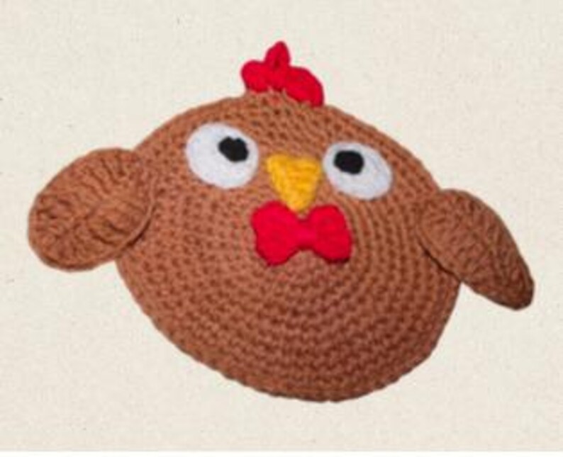 Puede incluir: Una cabeza de pollo de ganchillo hecha a mano, redonda y marr&oacute;n, con cresta y barbilla rojas, pico amarillo y ojos negros con borde blanco. El pollo tiene peque&ntilde;as alas redondeadas a cada lado. El fondo es de color beige claro.