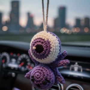 Puede incluir: Un adorno de coche tejido a ganchillo con forma de ojo morado y blanco con un pequeño cuerpo de pulpo morado. El adorno cuelga de una cuerda blanca, con un paisaje urbano borroso al fondo.