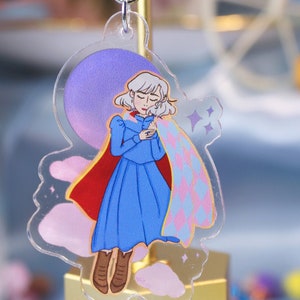Puede incluir: Un llavero de acrílico transparente con una ilustración de dibujos animados de una joven con cabello blanco que lleva un vestido azul y una capa a cuadros rosa y azul. La mujer está de pie sobre una nube con un fondo morado y rosa.