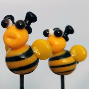 May include: Two small glass bee figurines with black and yellow stripes, black eyes, and a white dot on one bee's head. The bees are on metal rods.