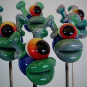 May include: A group of five glass alien figurines with green, blue, and red details. Each figurine has a single large eye and two small antennae. The figurines are mounted on metal pins.