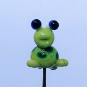May include: A green glass frog figurine with blue eyes and white pupils. The frog is sitting on a metal pin and has a spotted pattern.