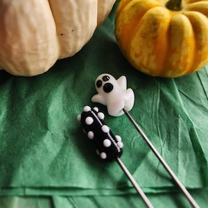 May include: Two Halloween-themed glass skewers. One is a white ghost with black eyes, and the other is a black cylinder with white polka dots. The skewers are on a green surface, with a white pumpkin and a small orange pumpkin in the background.