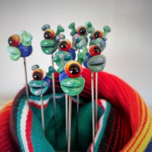 May include: A group of colorful glass figurines with googly eyes and green, blue, and red accents. The figurines are on metal skewers and are arranged in a striped red, white, and blue fabric.